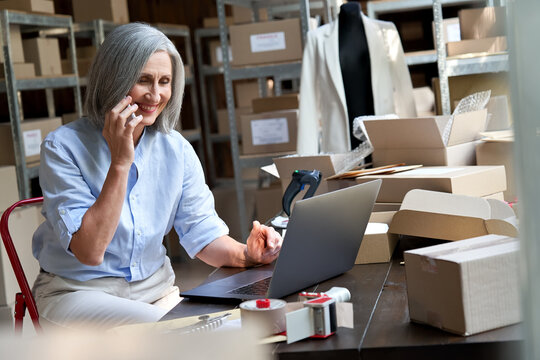 Older female fashion dropshipping business owner using laptop talking on phone in warehouse. Mature woman seller merchant checking online website order confirming ecommerce shipping parcel delivery.