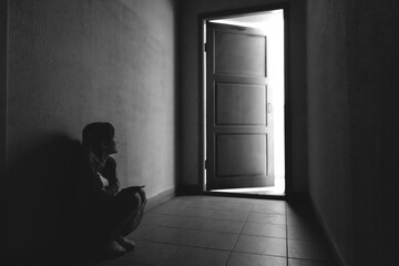 Little girl sitting in a dark corridor in front of the open door with light. Fear and...