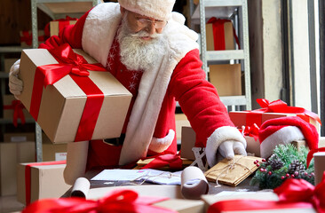 Old bearded Santa Claus, Saint Nicholas wearing costume holding gift box delivering presents preparing post shipping fast xmas holiday delivery standing at table in Merry Christmas workshop.