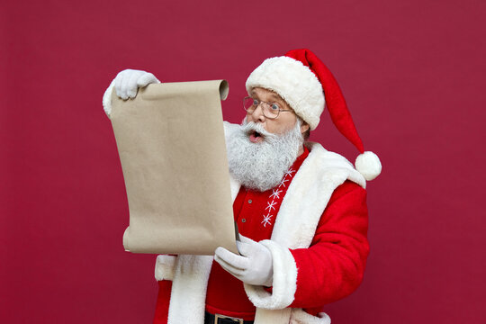 Shocked surprised old funny Santa Claus wearing costume holding parchment roll reading letter wish list preparing for Christmas holiday standing isolated on red background. Xmas wishlist concept.