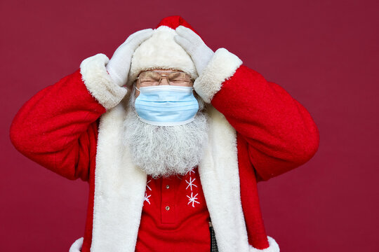Tired old bearded ill Santa Claus in panic wearing costume, face mask feeling headache, stress, getting sick on Christmas holidays standing on red background. Covid 19 coronavirus problem concept.