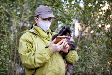 A woman wearing respirator mask and dialing a smartphone walking with a dog in the forest at COVID-19 period.