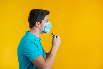 Young man in protective medical mask eating burger on yellow studio background. Guy makes fun, shows how to live during an epidemic.