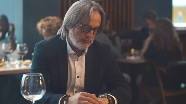 Portrait of aged man sitting in restaurant and checking time on wristwatch waiting for takeaway order