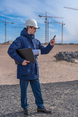 A construction worker in a white helmet with a laptop and documents under his arm is talking on the phone