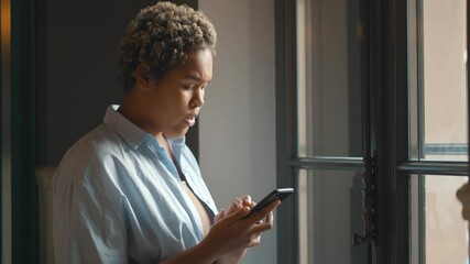 Anxious black lady reading depressing news on cellphone standing near window