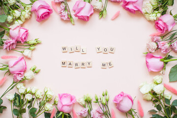 Inscription of wooden blocks will you marry me. Frame of delicate white and pink roses and eustomas on a light pink background. Layout.