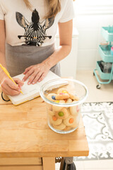 Obraz na płótnie Canvas Focused woman hands with cared nails writing a cookies recipe with a yellow pencil near a transparent jar full of colorful cookies.