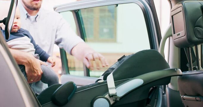Camera inside the car. Closeup baby car seat. Door opens and baby boy appears, carried by his smiling young bearded father, who puts the baby inside the baby car seat. Slow motion
