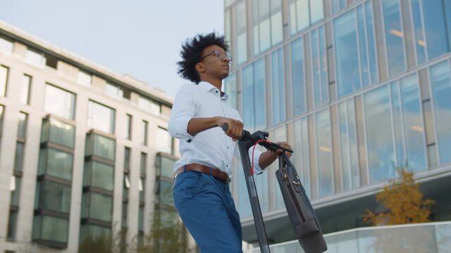African young businessman in glasses riding electro scooter near office building.