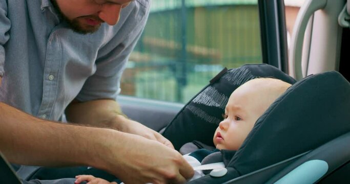 Camera inside the car. Closeup baby boy sits in the baby car seat inside of car. Young bearded father checks baby's fastened seat belts, smiles and talks to him, touching cheeks. Slow motion