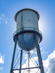 Old blue water tank placed on a tall structure with the sun behind and a blue sky with clouds in...