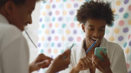 Afro man in bathrobe brushing teeth and using mobile phone in bathroom