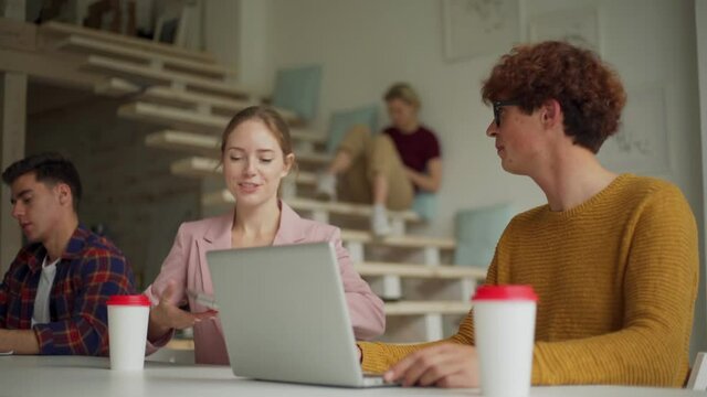 Tracking medium shot of young woman with laptop and takeaway coffee cup entering office after break, sitting down at desk and discussing project on laptop with male colleague in eyeglasses