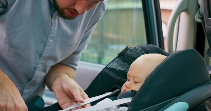 Camera inside the car. Closeup baby boy sits in the baby car seat inside of car. Young bearded father fastens seat belts, touches cheeks, and closes the door. Slow motion