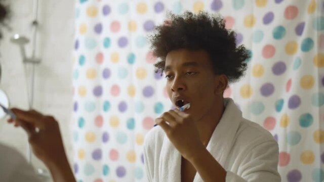 Afro man brushing teeth with toothbrush standing near mirror in bathroom