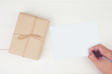 Hand of man writing letter with blank greeting card and having gift box with kraft paper on wooden table, present and package, postcard mockup, top view, flat lay, copy space, holiday concept.