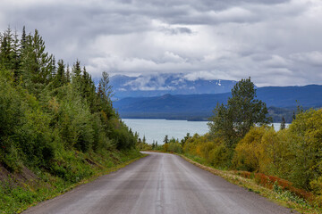 Fototapeta premium View of Scenic Road alongside Lake, surrounded by Trees and Mountains on a Cloudy Fall Day in Canadian Nature. Taken in Northern British Columbia, Canada.