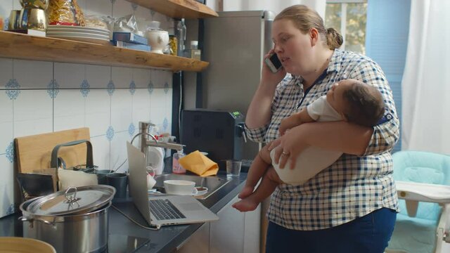 Young mother talking on phone holding sleeping baby at kitchen