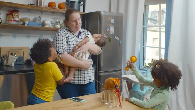 Tired white mother holding mixed race infant baby with toddler son and daughter playing in kitchen