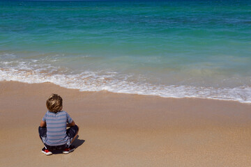 Boy crouching on the beach, facing the turquoise ocean with copy space on the right