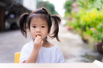 Adorable girl is eating oranges. It is difficult for children to eat fruit. Child wear white shirts. Little wooden table in front. Cute kid tied up with two black hair. Toddler are 3 years old.