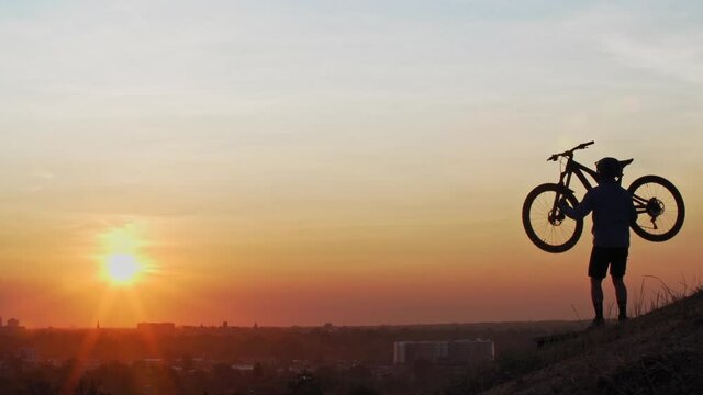 A male mountain biker, overlooking a town skyline on a hill, hoists his bike over his head as the low sun silhouettes his form.