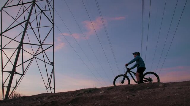 A male mountain biker, pulls under a power line tower to observe a dramatic setting, as the low sun silhouettes his form.  He stops and rests for a while to ponder.