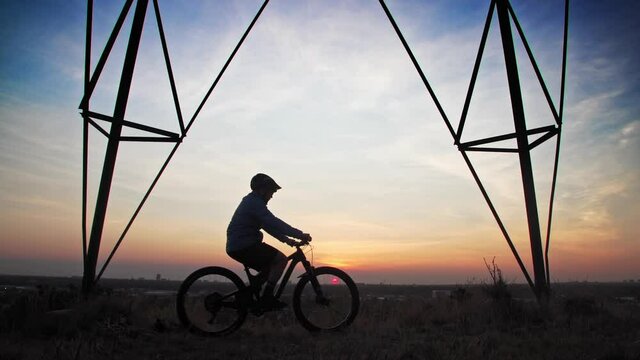 A male mountain biker, pulls under a power line tower to observe a town skyline, as the low sun silhouettes his form.  He stops and rests for a while to ponder.