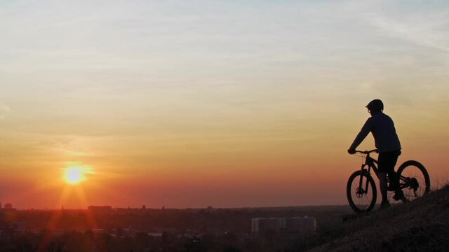 A male mountain biker, pulls up to a wood ramp atop a large hill to observe a town skyline, as the low sun silhouettes his form.  He stops and rests for a while to ponder.