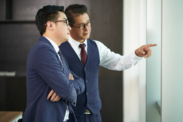 two asian business men standing by office window having a discussion