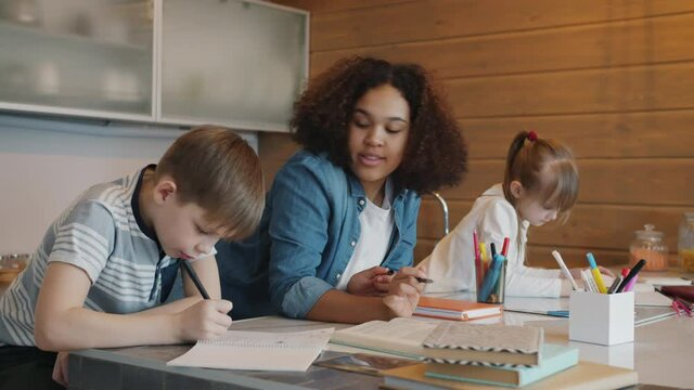 African American nanny tutor is helping diligent kids boy and girl with homework talking at table at home. Modern education and babysitting concept.