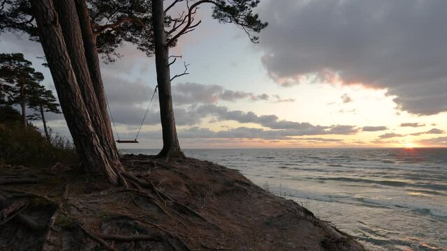 A wooden swing hanging between two pine trees growing on sandy hillside. Beautiful sunset on the Baltic sea at "Dutchman cap" place near Klaipeda.