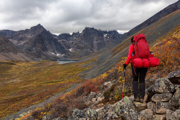 Fototapeta premium Woman Backpacking on Scenic Rocky Hiking Trail to Lake surrounded by Mountains during Fall in Canadian Nature. Taken in Tombstone Territorial Park, Yukon, Canada.