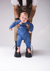 Mother encouraging baby kid To Take First Steps. A barefoot baby in a blue suit stands on mather's boots. Mom keeps safe  baby with his hands. 