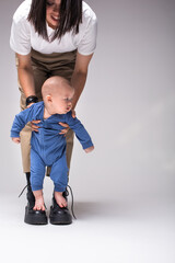 Mother encouraging baby kid To Take First Steps. A barefoot baby in a blue suit stands on mather's boots. Mom keeps safe  baby with his hands. Baby looks sideways