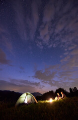 Silhouette of tourists spending time in the mountains in the evening near illuminated tent and a...