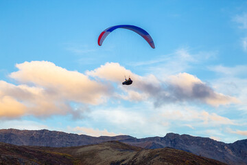 Fototapeta na wymiar Paraglider flying over Scenic Mountain Range, surrounded by Clouds in Canadian Nature. Taken in Tombstone Territorial Park, Yukon, Canada.