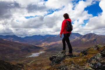 Fototapeta premium Woman Standing and Looking at Scenic Road and Mountain Range on a Cloudy Fall Day in Canadian Nature. Taken in Tombstone Territorial Park, Yukon, Canada.