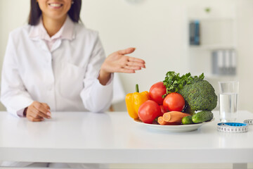 Smiling woman doctor pointing and recommending fresh vegetables for dieting during online consultation