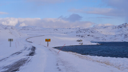 Icy road E69 with road sign displaying distance to Nordkapp (45km) near Honningsvåg, Norway, Scandinavia with red snowplow markings, remote house, snow-covered landscape and arctic sea in winter time.