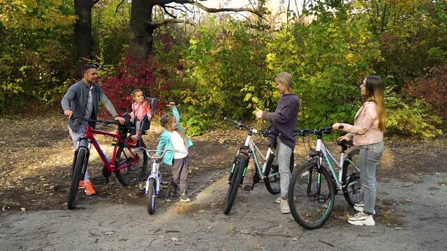 Family of five standing with bicycles in autumn park and showing thumbs up to each other, yellow and red trees on background. Parents and children feeling happy after biking outside