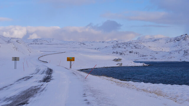 Icy road E69 with road sign displaying distance to Nordkapp (45km) near Honningsv&aring;g, Norway, Scandinavia with red snowplow markings, remote house, snow-covered landscape and arctic sea in winter time.