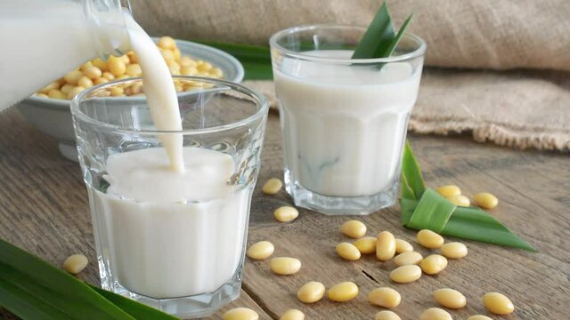 Pouring soy bean milk into glass with soybeans seed on wooden in the background.