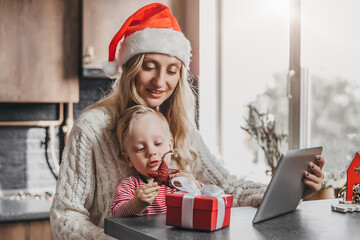 Family New Year and Christmas holidays. Mom with a little son in a festive Christmas hat are looking at toys and gifts and are sitting in the kitchen at the table against the background of the window