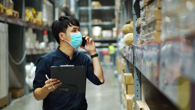 man worker with medical mask talking on a mobile phone and holding clipboard to checking inventory in the warehouse during coronavirus (covid-19) pandemic.