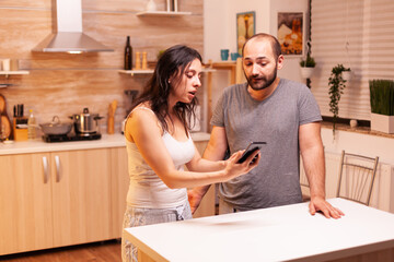 Wife screaming at unfaithful husband in the kitchen. Jealous woman cheated angry frustrated offended irritated accusing her man of infidelity showing him messages on smartphone yelling desperate.