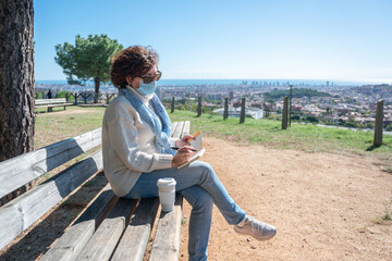 A woman a surgical mask, sitting on a bench writing in her notebook with the city in the background