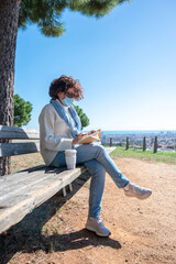 A woman a surgical mask, sitting on a bench writing in her notebook with the city in the background