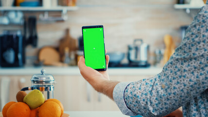 Mature man holding smartphone with green screen during breakfast in kitchen. Elderly person with...